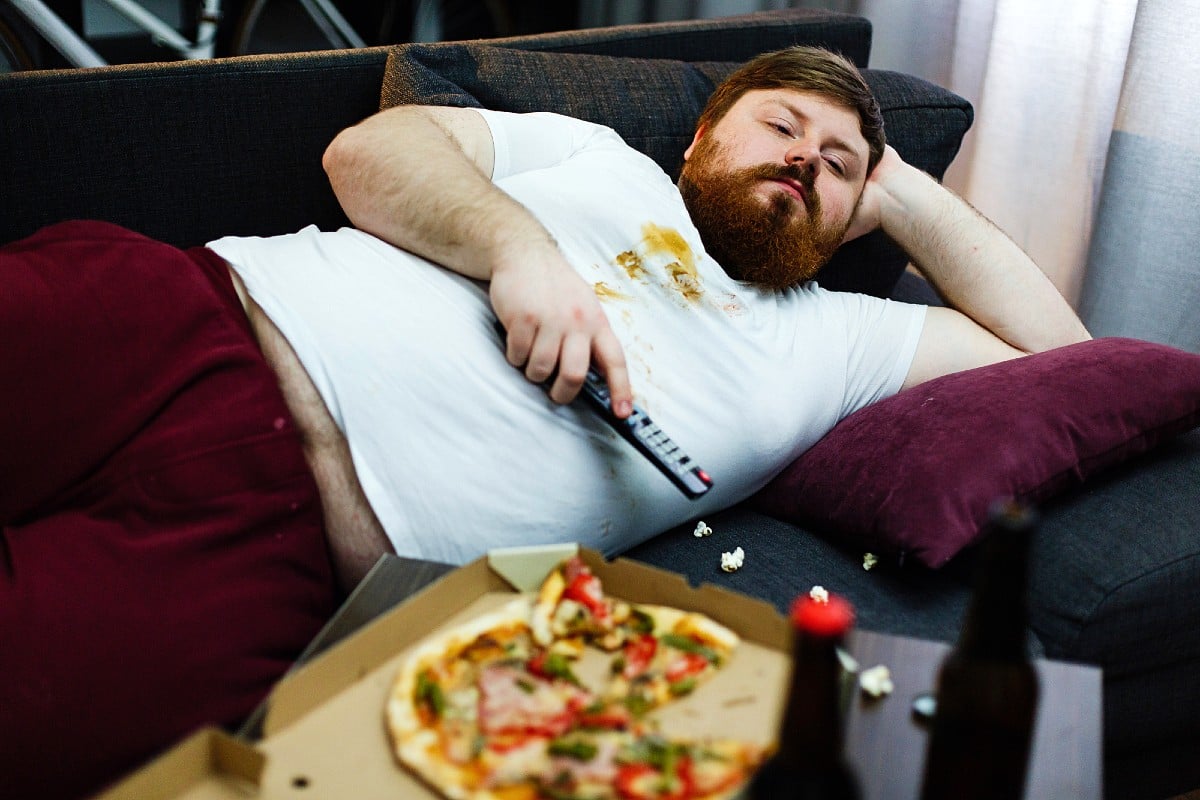 Lazy fat man eating pizza while lying on the couch watching TV